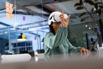 Working late at office, biracial businesswoman wearing VR headset, pointing upward