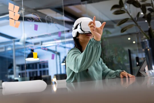 Working late at office, biracial businesswoman wearing VR headset, pointing upward