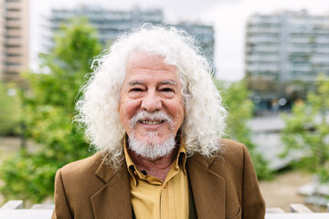 Happy senior man smiling at camera standing over urban background. Portrait of mature trendy male with white long hair and beard in trendy clothes outdoors.