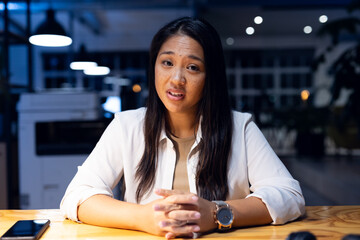 At office, biracial businesswoman sitting during video call, talking, working late