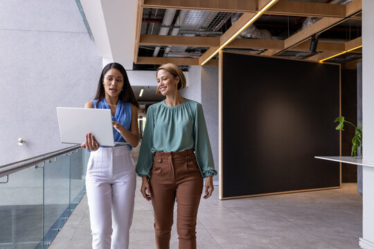 Diverse office team and biracial woman in blue talk laptop content while walking, copy space