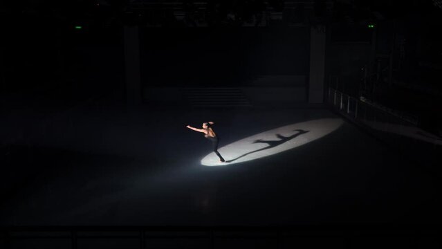 Girl skater in black overalls on dark rink performs turn, Axel jump and four spins in balance, grab skate blade of free leg behind her back and makes three Biellmann spins. Shadow image in spotlight