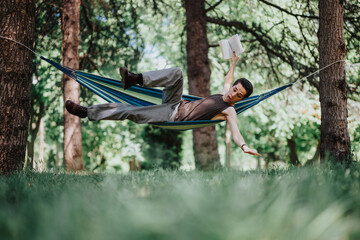 A joyful young man balances in a hammock while reading a book, exhibiting balance and relaxation...