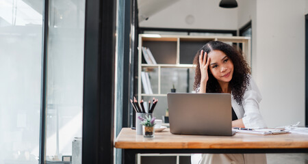Contemplative businesswoman with a hand on her forehead sits at her laptop in a sunlit, contemporary office space.