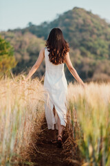 Back view of a woman in a flowing white dress walking down a narrow path in a lush wheat field, with a gentle mountain backdrop.