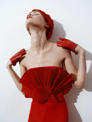 Elegant woman in a red dress and gloves posing with hands on head against white wall
