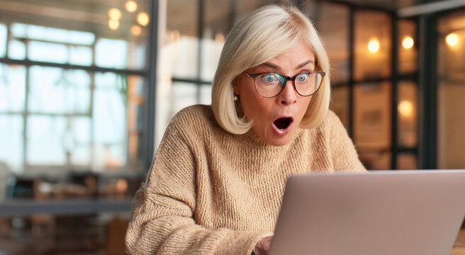 Mature woman with blonde hair and glasses has shocked expression, looking at screen of her laptop computer.