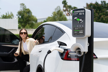 Young woman recharge her EV electric vehicle at green city park parking lot. Urban sustainability...