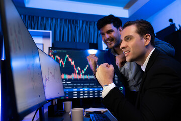 Cheerful two stock exchange traders raising fist up for digital currency achievement focusing on dynamic data background. Successful investors earning high profit analyzed by market graph. Sellable.