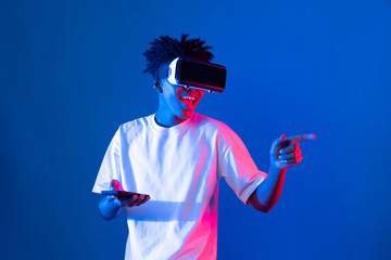 Young African American player wearing VR with using smartphone playing game hologram on blue pink...
