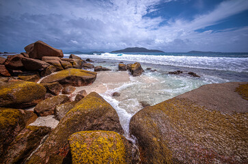 Waves are flattening around granite rocks on the Seychelles Islands. Sunny sky with white couds, and islands are on the horizon.