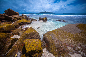 Large granite rocks are on the beach, with shallow water floating around them. The sky is sunny with white clouds, and island silhouettes are visible on the distant horizon.
