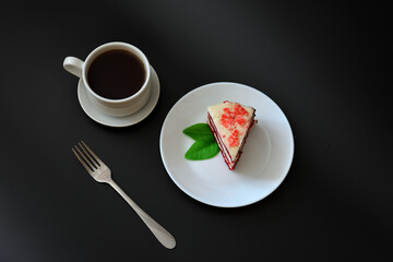 A piece of red velvet cake with mint, a fork and a cup of hot black coffee on a black background.