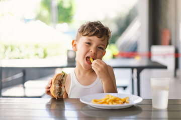 Little Boy Eating Sandwich and French Fries at Table. He appears focused on his meal, with a...