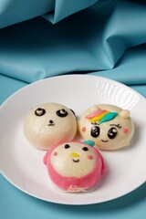 A whimsical treat: white steamed bun filled with chocolate, topped with a playful face complete with eyes, nose, mouth, and a hint of hair. Presented on a white plate against a blue backdrop.