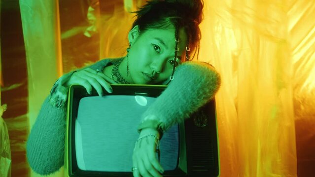 Beautiful Asian fashion model leaning on retro TV with no signal and posing at camera in studio with colorful illumination and plastic wrap background. Video portrait, zoom away shot