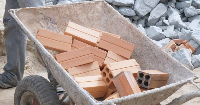 close up of a worker placing new, reddish brown bricks into a rusty metal wheelbarrow. 