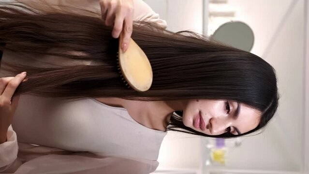 Vertical video. Strands brushing. Feminine routine. Beautiful woman in elegant blouse combing gorgeous healthy smooth long brunette hair in light home interior.
