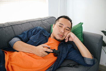 Bored man holding joystick, playing video games while lying on sofa at home