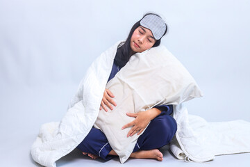 Young Asian woman in pajamas and sleep eye mask holding pillow falling asleep while covered with...