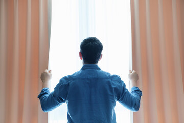 Young man in blue shirt looks out the window and opens the curtains, rear view.