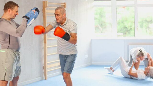 Coach and senior man pupil are engaged during training, boxing classes. Male trainer holds makiwara and helps student to work out force of blow, to box quickly and effectively