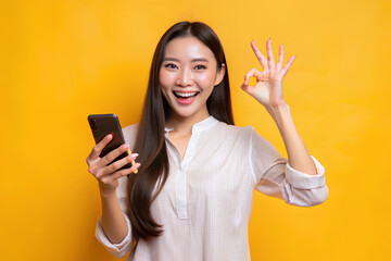 Cheerful beautiful Asian woman holding smartphone and shows ok sign on light yellow background