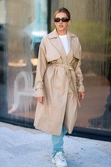 Fototapeta premium Coat. Outdoor lifestyle portrait of pretty young fashion model in urban background. Outdoor elegant fashionable woman walking in street. Summer lifestyle portrait of stylish girl wearing trendy outfit