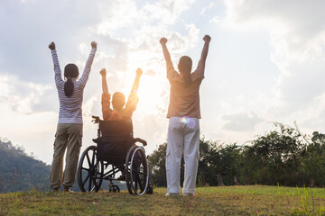 Rear view joyful family group raised hands with elderly women in wheelchair on mountain sunset...
