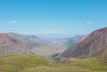 Sunlit wide long valley with green hills and river between multicolor mountains in sunny day. Vivid...
