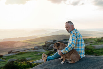 senior mature bearded man with red english bulldog sitting on top of mountain	