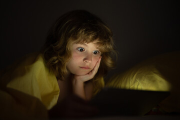 Kid is addicted to tablet, kid use tablet, watching cartoon. Child watching his tablet in the bed. Illuminated child face from device screen. Boy under the covers hold a tablet. Night time.