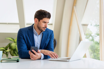 Confident businessman sitting at desk and using laptop and smartphone for work