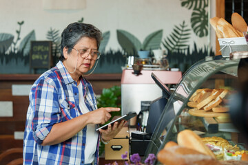 Elder asian barista in checked shirt attentively manages cafe affairs on tablet, artisan breads...