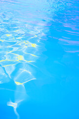 Water surface in swimming pool with bright blue color. Background of pool surface in the morning with sunlight. The concept of sun reflection, summer, and relaxation. Vertical, rippled water texture.