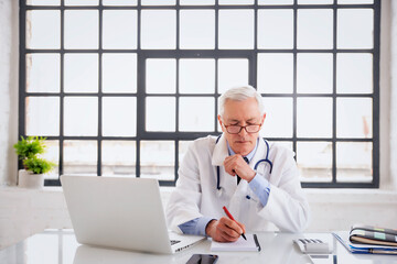 Elderly male doctor sitting in doctor's room and using laptop