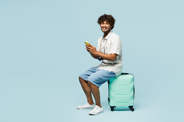 Full body traveler Indian man wears white casual clothes sit on bag using mobile cell phone...