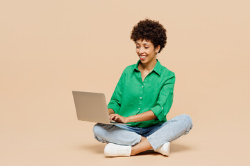 Full body happy young IT woman of African American ethnicity wear green shirt casual clothes sit hold use work on laptop pc computer isolated on plain pastel light beige background. Lifestyle concept.