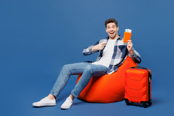 Traveler man wear casual clothes sit in bag chair near suitcase hold point finger on passport...
