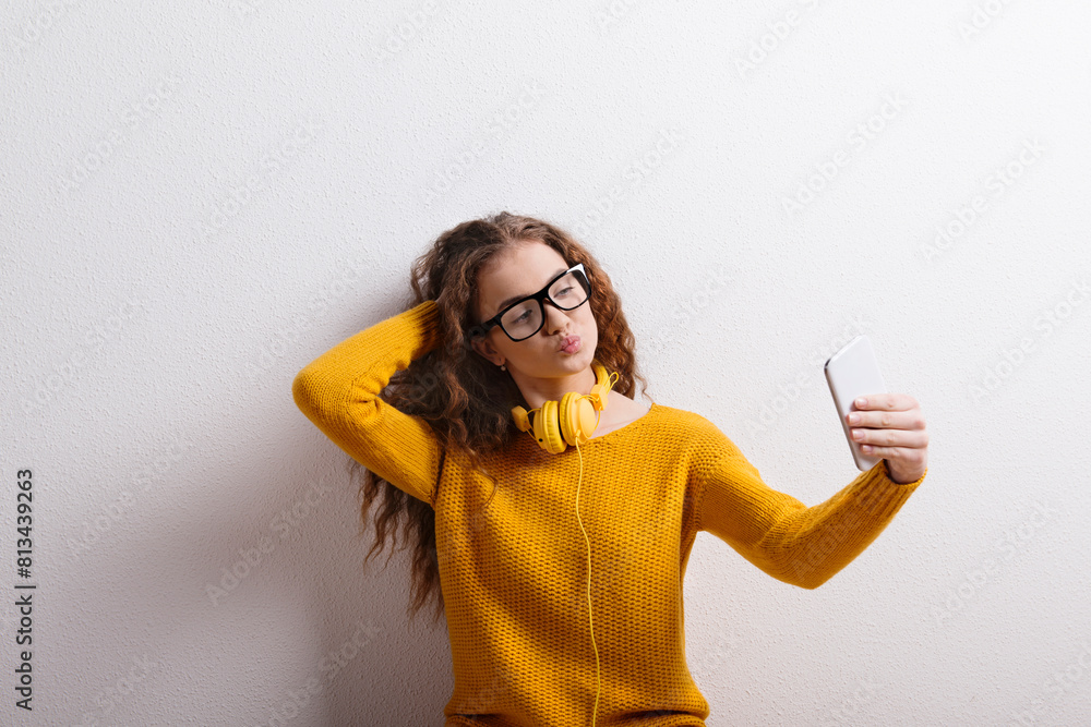 Wall mural portrait of a gorgeous teenage girl with curly hair and headphones,taking selfie. studio shot, white