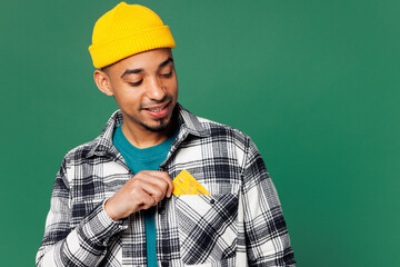 Young man of African American ethnicity he wears shirt blue t-shirt yellow hat hold put mock up of credit bank card into pocket isolated on plain green background studio portrait. Lifestyle concept.