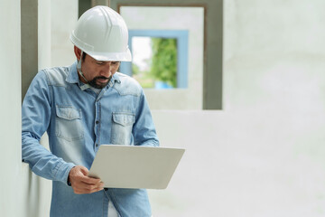 Man in blue denim shirt and white safety helmet studies laptop screen, concentration visible,...