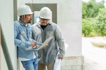 In construction setting, two focused men, helmets on, discuss over laptop, with one typing,...