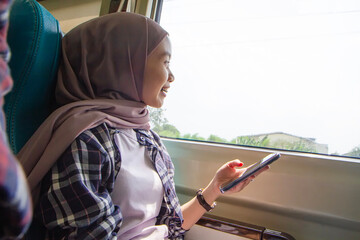 happy asian muslim woman holding her mobile phone while sitting beside big window train wagon. traveling concept