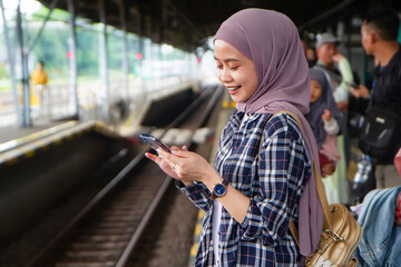 happy asian muslim woman holding phone standing on train station platform waiting for the train....