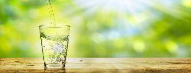 pouring mineral water in a transparent water glass isolated on wooden plank in front of blurred nature bokeh background outdoors with copy space © winyu