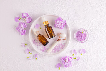 Two Dropper Bottles With Natural Skin Care Product, Oil For Massage, Stone Roller Massager On White Round Tray Among Flowers. Top view