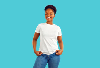 Studio portrait of happy woman in casual clothes. Beautiful confident young African American girl in blue jeans and white mockup T shirt standing isolated on turquoise background. Fashion concept