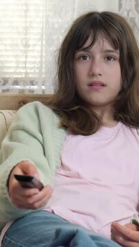 Child, teenage is sitting on the couch at home, using the remote control to switch channels on the TV.