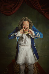 Hungry elderly man, dressed as king in classical baroque style clothes eating modern hamburger...